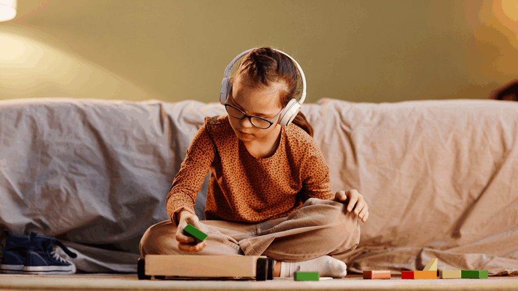 Junges Mädchen mit Brille und Kopfhörern sitzt auf dem Boden vor einer Couch und sortiert bunte Holzklötze – ruhige Spielumgebung, mögliches Symbol für Autismus, sensorische Integration oder therapeutische Förderung im Kindesalter.