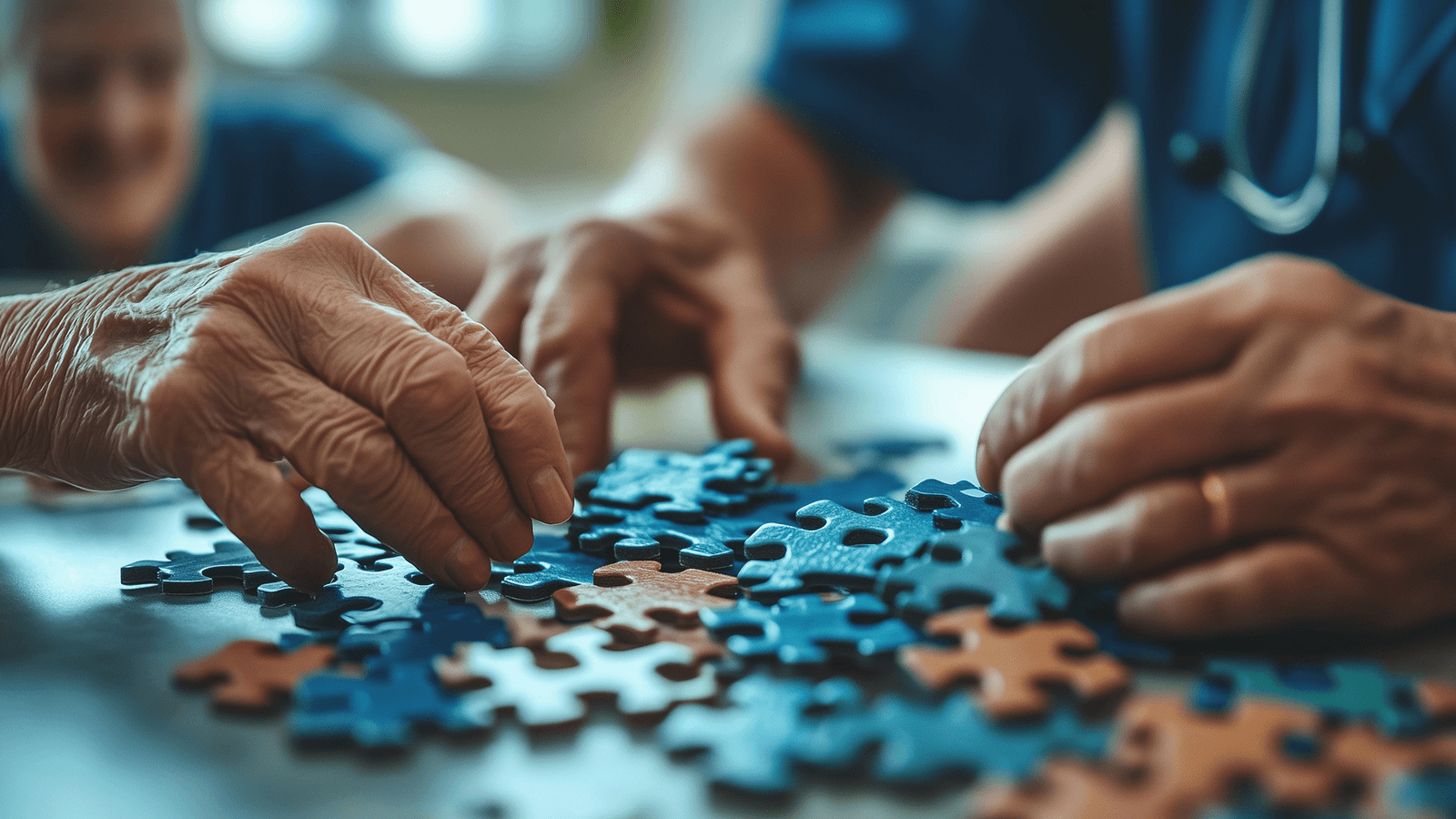 Zwei ältere Personen legen gemeinsam ein Puzzle – Nahaufnahme der Hände, im Hintergrund eine Pflegekraft in blauer Kleidung. Symbol für Gedächtnistraining, Feinmotorik-Übungen oder ergotherapeutische Förderung im Alter.