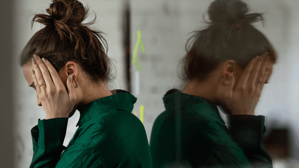 Junge Frau lehnt sich mit den Händen an den Kopf gegen eine Glaswand – ihr Spiegelbild zeigt dieselbe Haltung. Die Szene vermittelt emotionale Erschöpfung oder psychische Belastung, wie sie etwa in einer seelischen Krise auftreten kann. Thema: Psychische Gesundheit und ergotherapeutische Unterstützung.