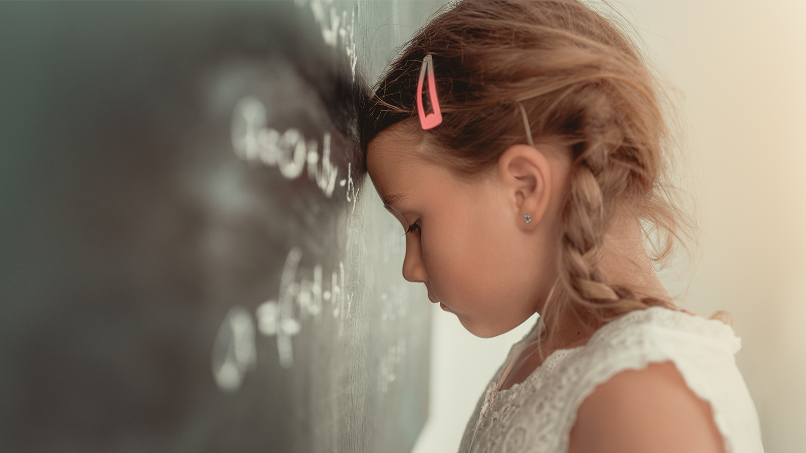 Ein junges Mädchen mit ADHS und zwei geflochtenen Zöpfen lehnt mit geschlossenen Augen und der Stirn gegen eine Schultafel. Sie wirkt erschöpft oder überfordert. Auf der Tafel sind verschwommene mathematische Formeln zu erkennen.