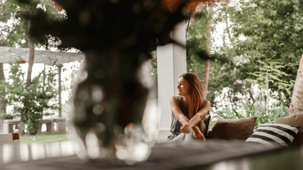 Eine junge Frau sitzt entspannt auf einer Terrasse im Grünen, blickt lächelnd zur Seite und genießt den Blick in die Natur. Im Vordergrund unscharfe Pflanzen, im Hintergrund Bäume und Sträucher. Symbol für die heilende Kraft der Natur und den Einsatz von Natur- und Gartentherapie in der Ergotherapie zur Förderung von Wohlbefinden und seelischer Gesundheit.