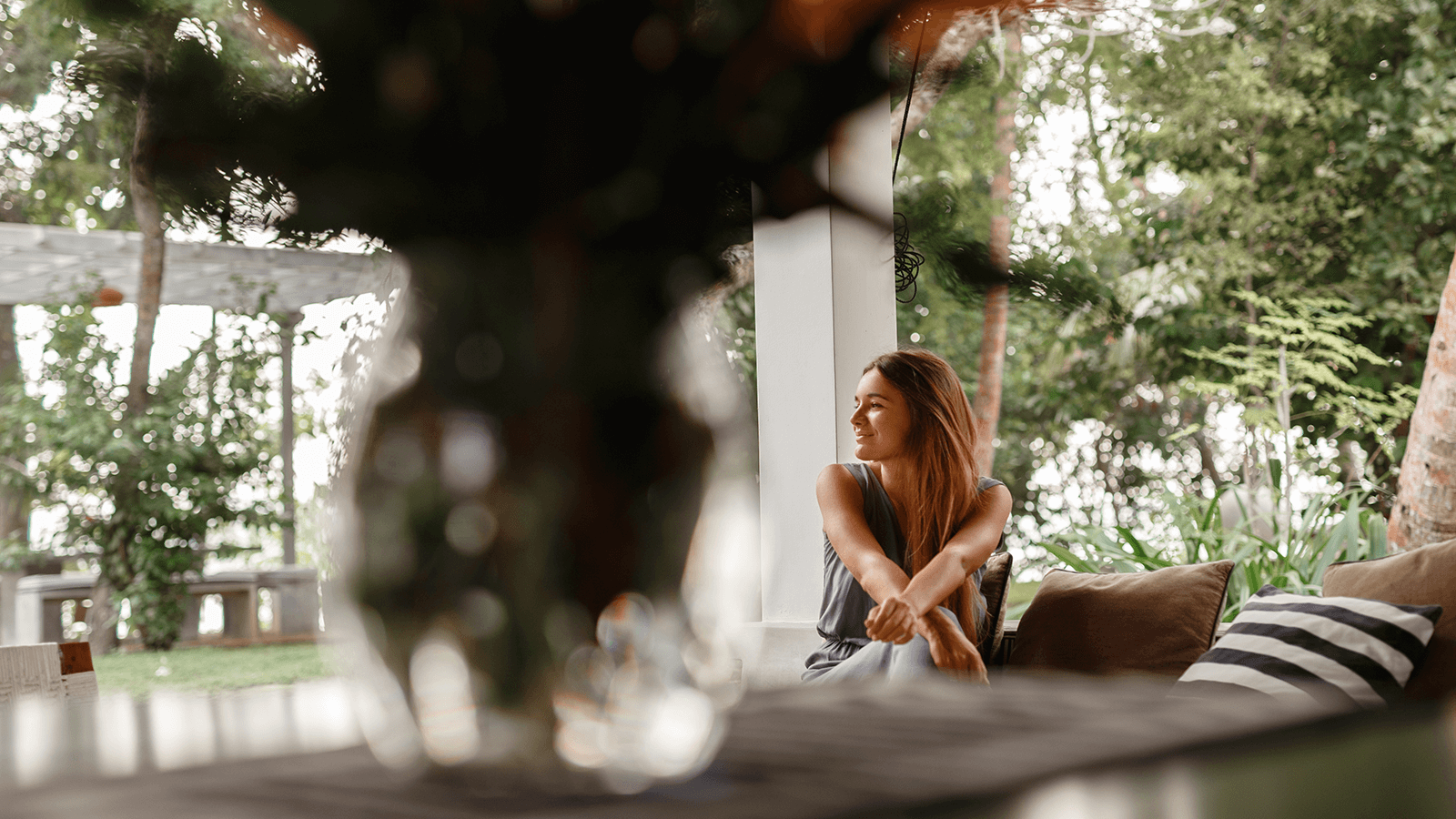 Eine junge Frau sitzt entspannt auf einer Terrasse im Grünen, blickt lächelnd zur Seite und genießt den Blick in die Natur. Im Vordergrund unscharfe Pflanzen, im Hintergrund Bäume und Sträucher. Symbol für die heilende Kraft der Natur und den Einsatz von Natur- und Gartentherapie in der Ergotherapie zur Förderung von Wohlbefinden und seelischer Gesundheit.