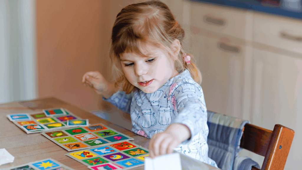 Kleines Mädchen sitzt am Tisch und spielt konzentriert mit bunten Bildkarten, die verschiedene Motive in kräftigen Farben zeigen – eine Übung aus der Ergotherapie für Kinder, bei der Töne und Farben zur Förderung der Wahrnehmung eingesetzt werden.