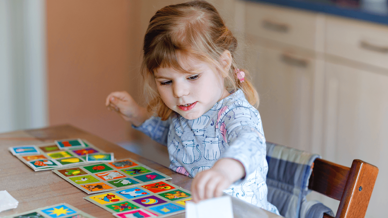 Kleines Mädchen sitzt am Tisch und spielt konzentriert mit bunten Bildkarten, die verschiedene Motive in kräftigen Farben zeigen – eine Übung aus der Ergotherapie für Kinder, bei der Töne und Farben zur Förderung der Wahrnehmung eingesetzt werden.