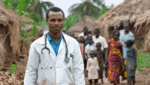 Ein afrikanischer Arzt in weißem Kittel mit Stethoskop um den Hals steht im Vordergrund, leicht lächelnd und direkt in die Kamera blickend. Im Hintergrund gehen mehrere Dorfbewohner, darunter Frauen und Kinder in bunter Kleidung, auf einem Weg zwischen strohgedeckten Hütten entlang. Die Szene vermittelt Hoffnung und gemeinschaftlichen Einsatz für medizinische Versorgung in ländlichen Regionen Afrikas.