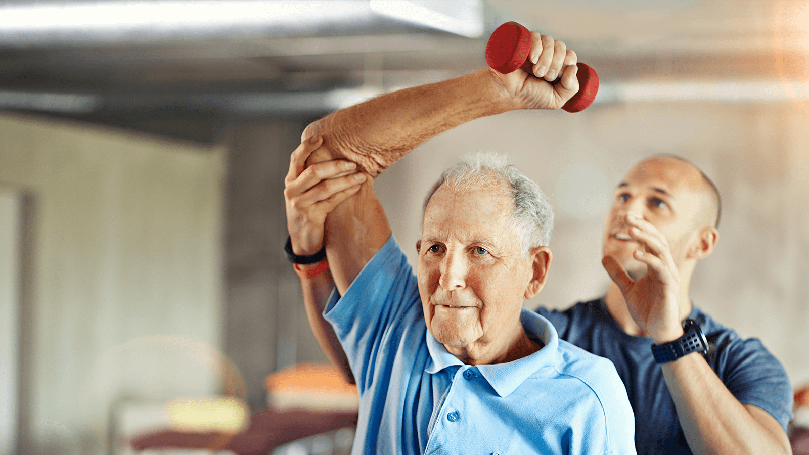 Ein älterer Mann in einem blauen Poloshirt hebt mit Unterstützung eines Therapeuten einen roten Hantelarm. Zeigt gezieltes Alltagstraining zur Erhaltung der Beweglichkeit.