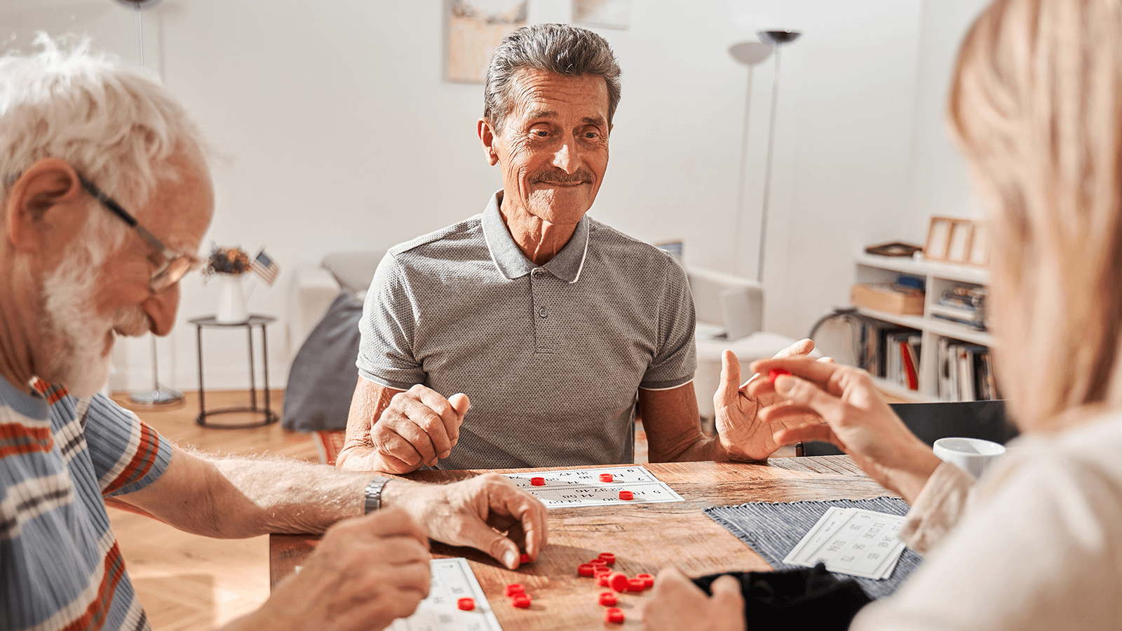 Drei ältere Menschen sitzen gemeinsam an einem Tisch und spielen ein Brettspiel mit roten Spielsteinen. Alle wirken konzentriert und zufrieden, der Mann in der Mitte lächelt leicht. Die Umgebung wirkt wohnlich und hell, mit Bücherregal und Tischdeko im Hintergrund. Das Bild vermittelt Gemeinschaft, Lebensfreude und Aktivität im Alter.