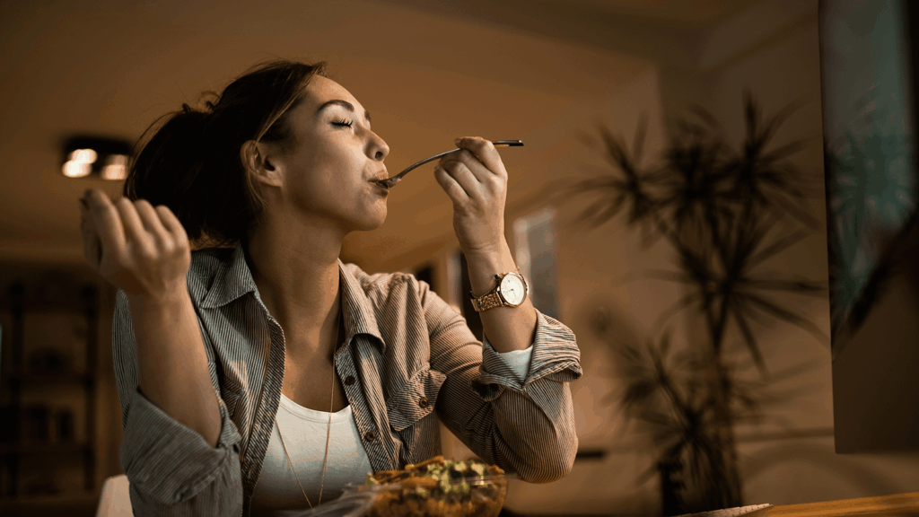 Junge Frau sitzt abends entspannt am Tisch und genießt bewusst eine Mahlzeit, während sie genüsslich einen Löffel zum Mund führt – Symbol für gesunde Ernährung als unterstützenden Teil der Rehabilitation.