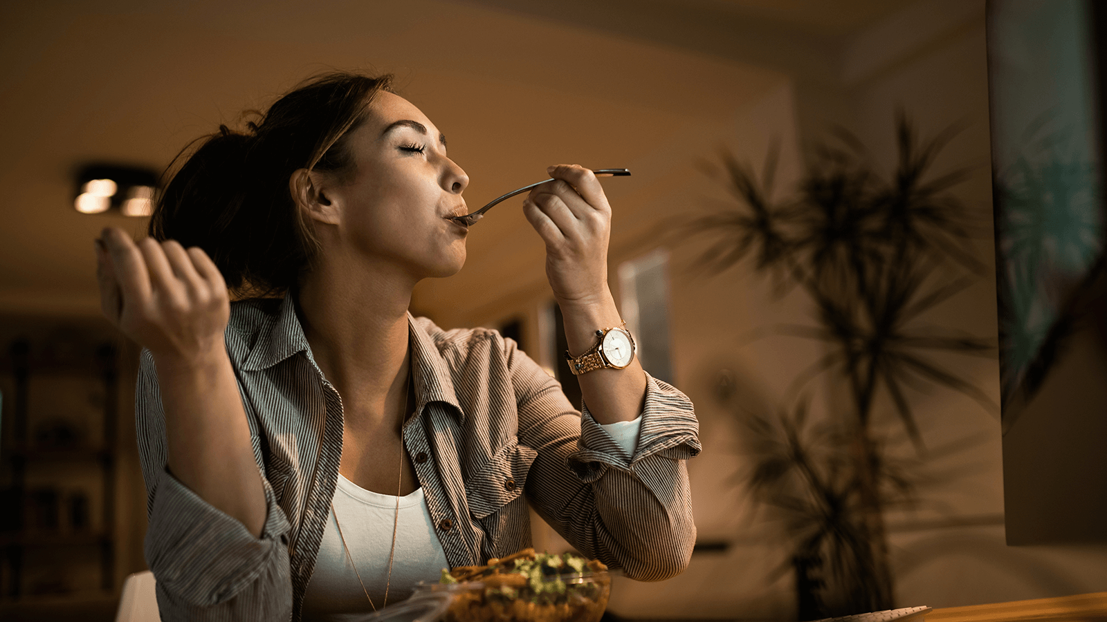 Junge Frau sitzt abends entspannt am Tisch und genießt bewusst eine Mahlzeit, während sie genüsslich einen Löffel zum Mund führt – Symbol für gesunde Ernährung als unterstützenden Teil der Rehabilitation.