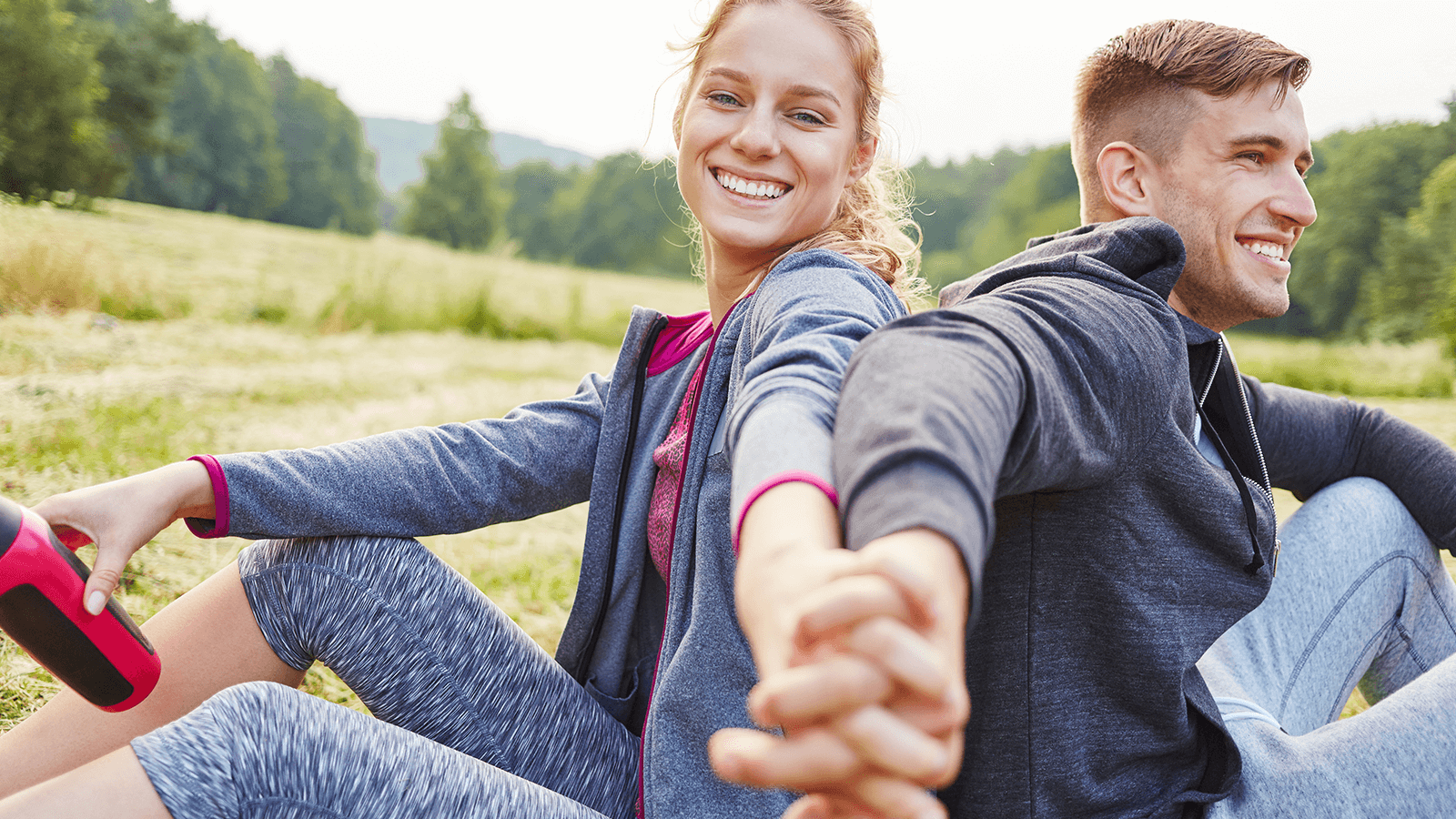 Junge Frau und junger Mann sitzen Rücken an Rücken auf einer Wiese, halten sich an den Händen und lächeln entspannt – Symbol für Vitalität, Lebensfreude und gemeinsame Bewegung an der frischen Luft.