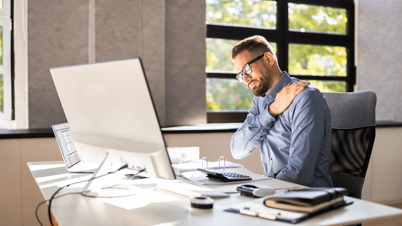 Mann im Büro sitzt vor dem Computer und fasst sich mit schmerzverzerrtem Gesicht an die Schulter – Symbol für Verspannungen, Überlastung oder Fehlhaltung durch langes Sitzen am Bildschirmarbeitsplatz.