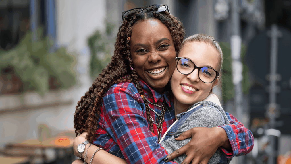 Zwei Frauen unterschiedlicher Herkunft umarmen sich herzlich und lächeln in die Kamera. Die Frau links trägt ein kariertes Hemd in Rot und Blau und hat lange, gelockte Haare, die Frau rechts trägt eine Brille, einen grauen Hoodie und hat ihre Haare zu einem Pferdeschwanz gebunden. Die Szene spielt im Freien, unscharfer Hintergrund mit städtischem Flair. Das Bild steht für interkulturelle Kompetenz, Herzlichkeit und gegenseitiges Verständnis.