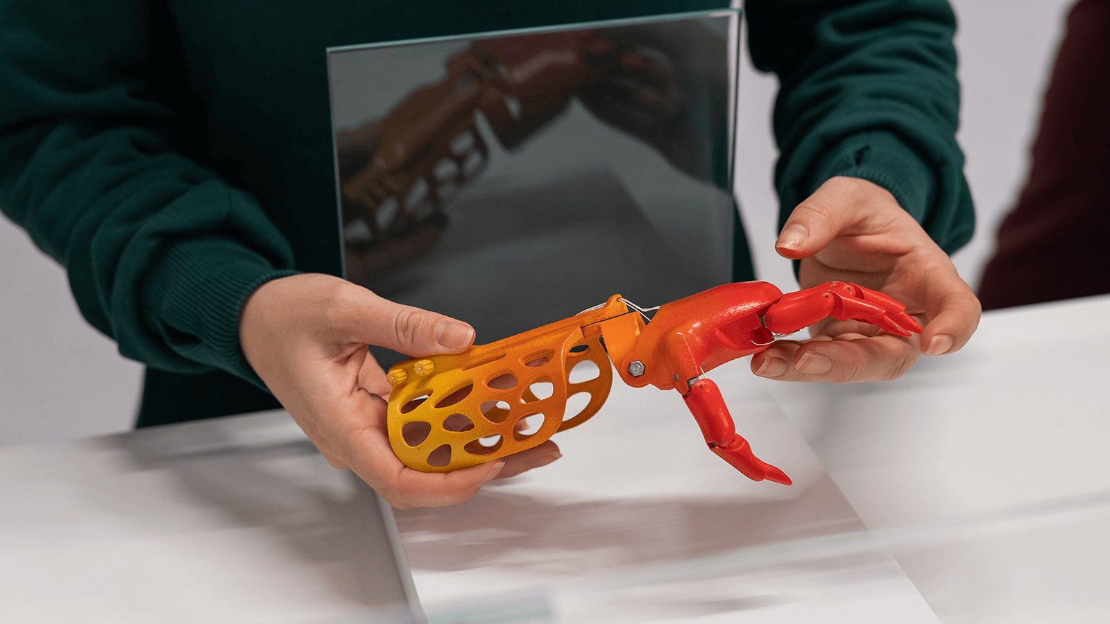Person hält mit beiden Händen eine bunte, orange-rote Handprothese aus dem 3D-Drucker. Die Prothese hat ein offenes, gitterartiges Design. Im Hintergrund spiegelt sich das Objekt in einer Glasscheibe.