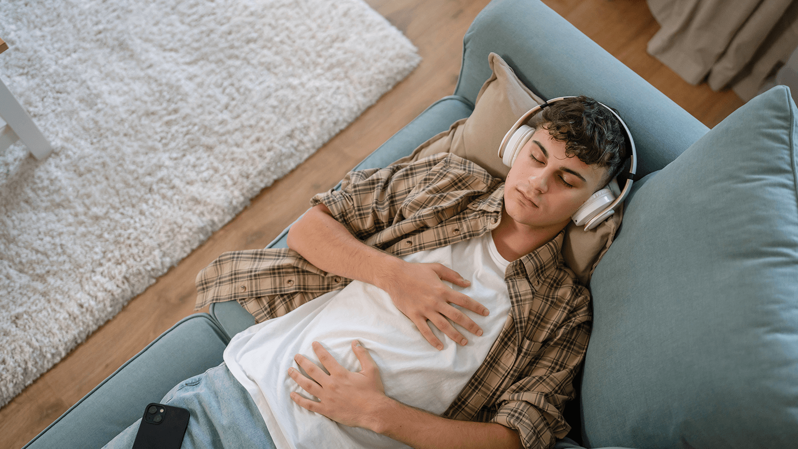 Junger Mann liegt entspannt auf einem hellblauen Sofa, trägt große weiße Kopfhörer und hat die Augen geschlossen. Er hält beide Hände locker auf seinem Bauch, während neben ihm ein Smartphone liegt. Die Szene vermittelt Ruhe, Achtsamkeit und Entspannung im Alltag zur Stressreduktion.