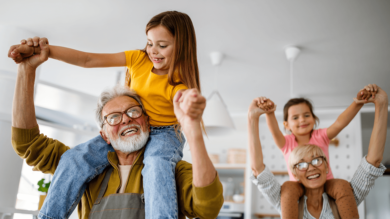 Zwei Großeltern spielen lachend mit ihren Enkelkindern zu Hause. Ein älterer Mann trägt ein Mädchen auf den Schultern, während eine ältere Frau im Hintergrund ebenfalls ein Kind auf den Schultern hält. Alle wirken fröhlich und energiegeladen in einem hellen, freundlichen Wohnraum.