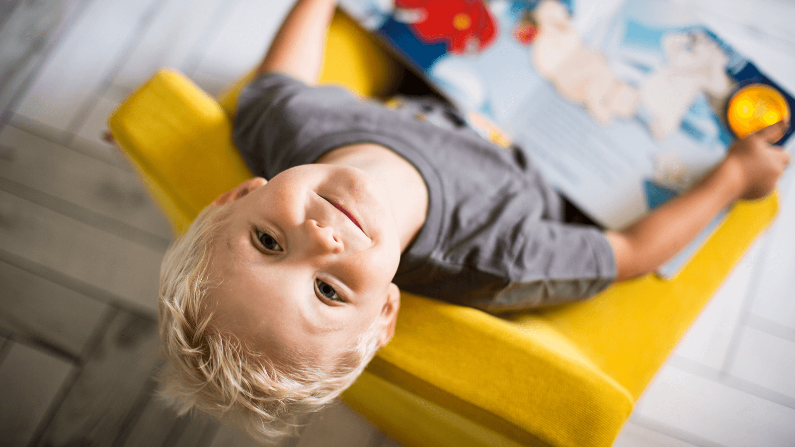 Ein kleiner blonder Junge liegt rücklings in einem leuchtend gelben Stuhl, schaut lachend zur Kamera und hält ein aufgeschlagenes Kinderbuch in den Händen.