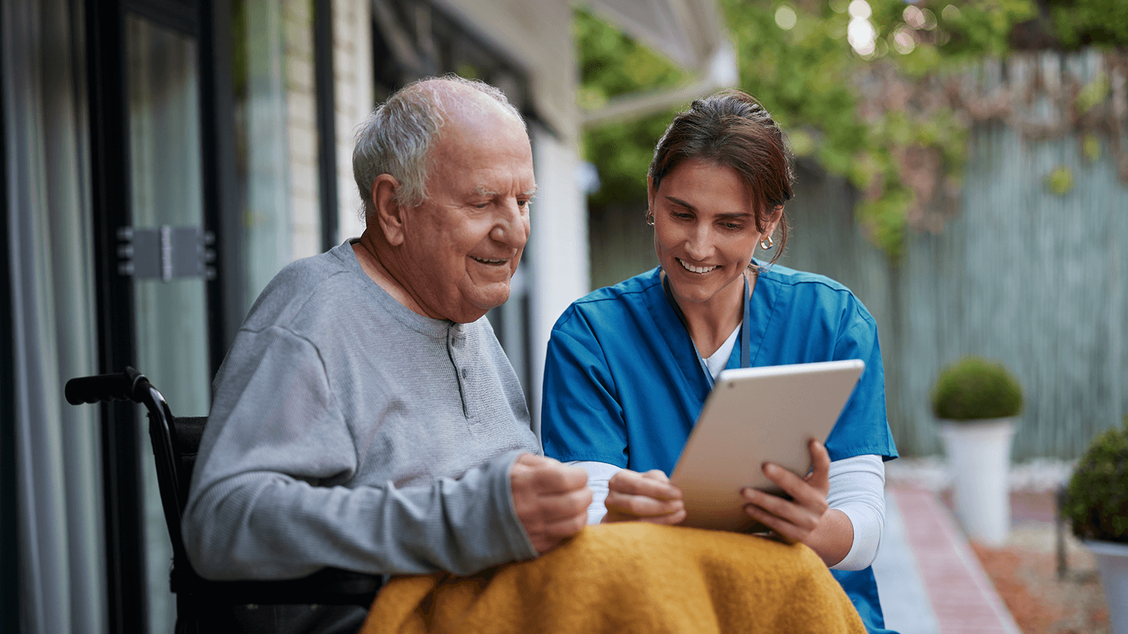 Ein älterer Mann im Rollstuhl sitzt im Freien neben einer Pflegekraft, beide lächeln und schauen gemeinsam auf ein Tablet. Symbolisiert Teletherapie und Betreuung zu Hause.