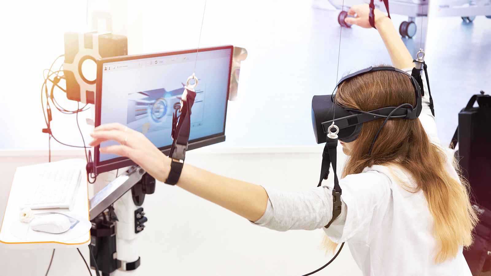 Eine junge Frau trägt eine Virtual-Reality-Brille und Armaufhängungen, während sie vor einem Bildschirm interaktive Übungen macht. Darstellung moderner, technikgestützter Rehabilitation.