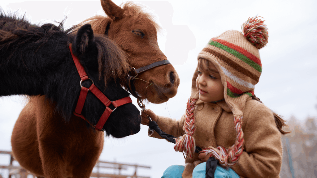 Ein junges Mädchen in warmer Winterkleidung mit gestreifter Bommelmütze streichelt und führt zwei kleine Ponys, die freundlich und ruhig wirken. Die Szene vermittelt Nähe, Vertrauen und Hoffnung, wie sie in tiergestützter Ergotherapie entstehen kann.