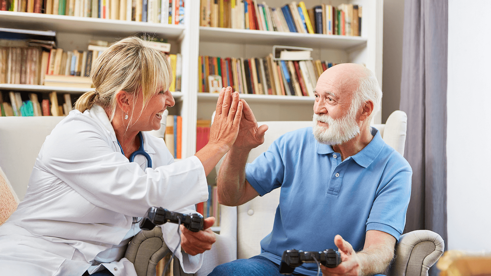 Ärztin in weißem Kittel sitzt neben einem älteren Mann mit grauem Bart. Beide halten Spielecontroller in den Händen und lachen sich an, während sie sich zur Begrüßung oder zum Erfolgserlebnis mit einer High-Five-Geste abklatschen. Spielerische Therapie.